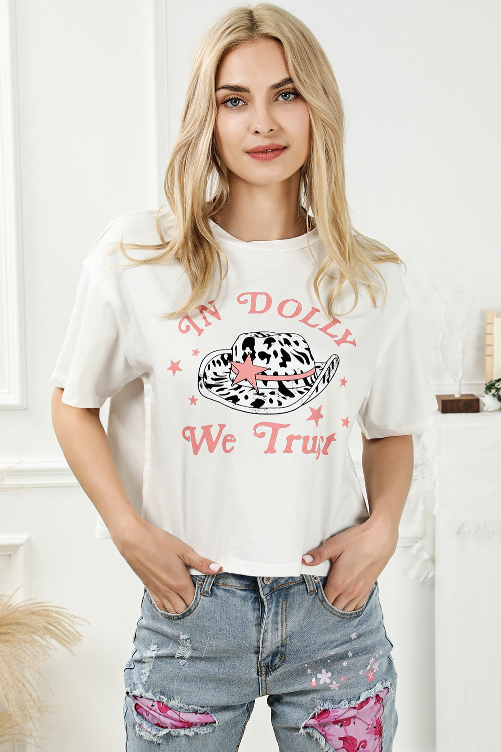 Woman wearing IN DOLLY WE TRUST round neck T-shirt, white with pink lettering, paired with denim jeans.